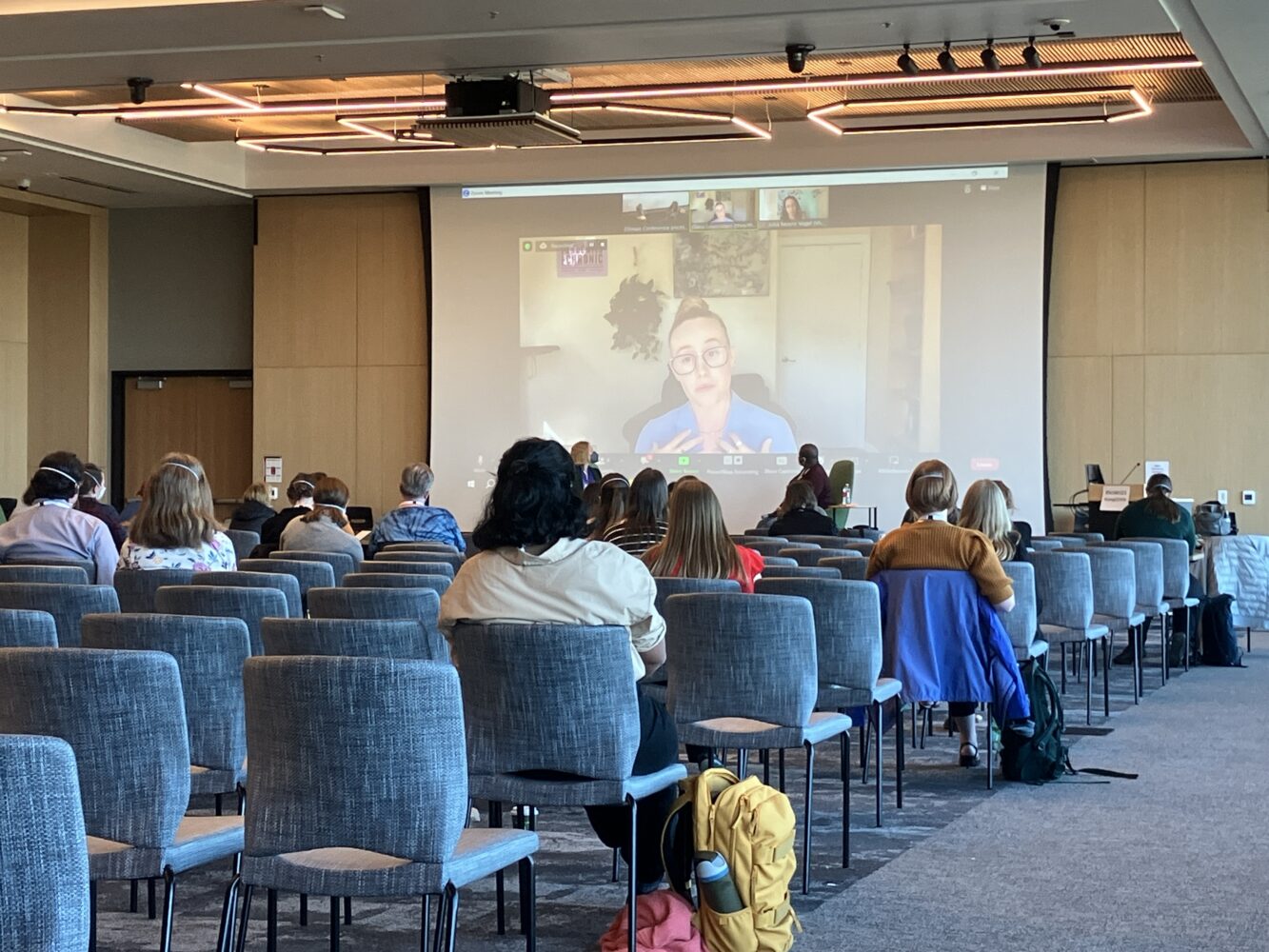 Conference attendees sit in chairs to watch a hybrid session on covering Long COVID. A panelist is on a video call on a projector screen at the front of the room.