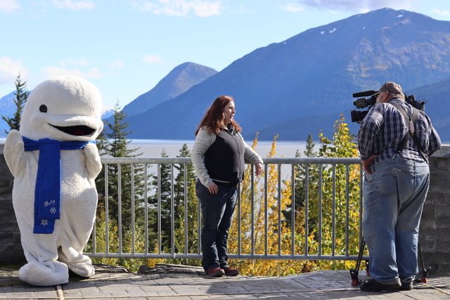 A camera crew interviewing a woman standing next to railing with water and mountains in the background. A person dressed up as beluga whale stands next to the woman.