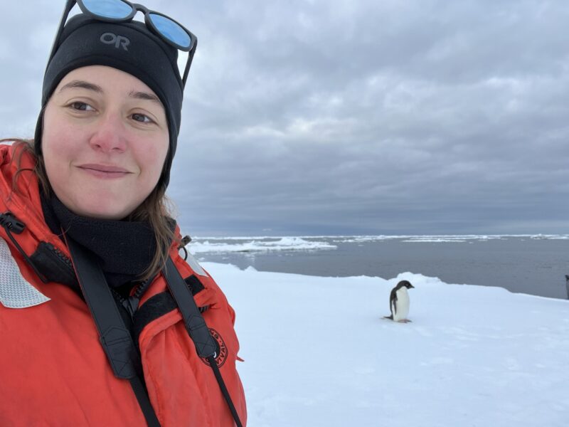 A woman dressed in orange safety gear takes a selfie with a penguin in a snowy landscape. 